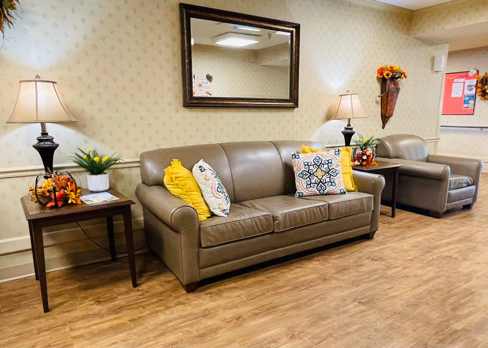 A cozy seating area in a senior living facility hallway featuring a brown leather sofa and matching armchair, both adorned with decorative yellow and patterned pillows. Two side tables with table lamps, floral arrangements, and autumn-themed decorations flank the seating. A large rectangular mirror hangs on the wall above the sofa, reflecting part of the ceiling and hallway.