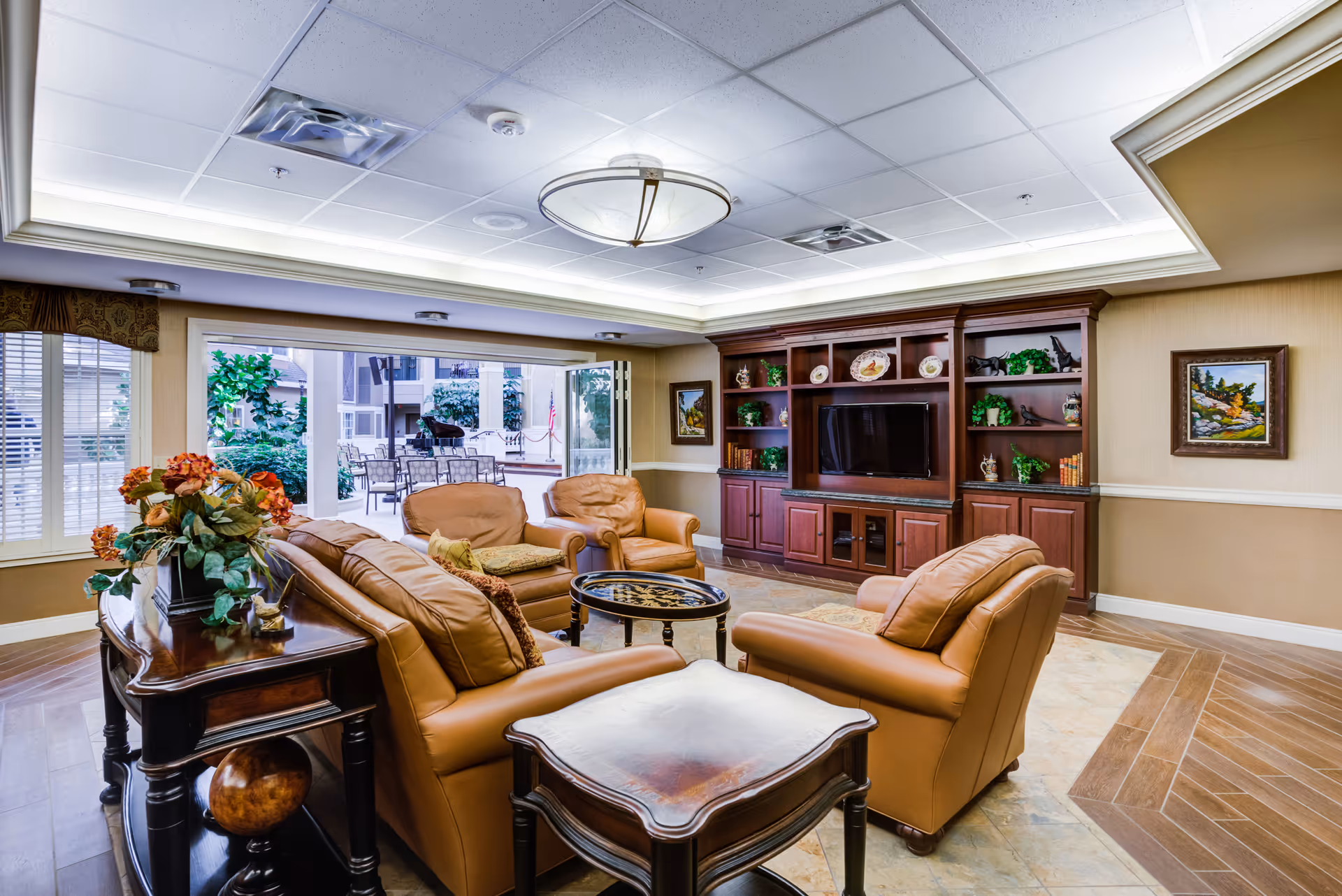 A cozy living room area with tan leather armchairs and a sofa arranged around a small round coffee table. Behind the seating is a wooden console table with a floral arrangement. The room features a large wooden entertainment center with a flat-screen TV, decorative plates, books, and plants. Large windows and an open sliding door reveal an outdoor patio area with tables and chairs. The ceiling has recessed lighting and a central light fixture.