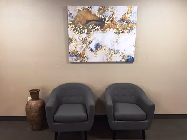 Two gray upholstered armchairs placed side by side against a beige wall with a large abstract painting featuring gold, white, and blue colors hanging above them. To the left of the chairs is a tall decorative vase with a rustic finish.