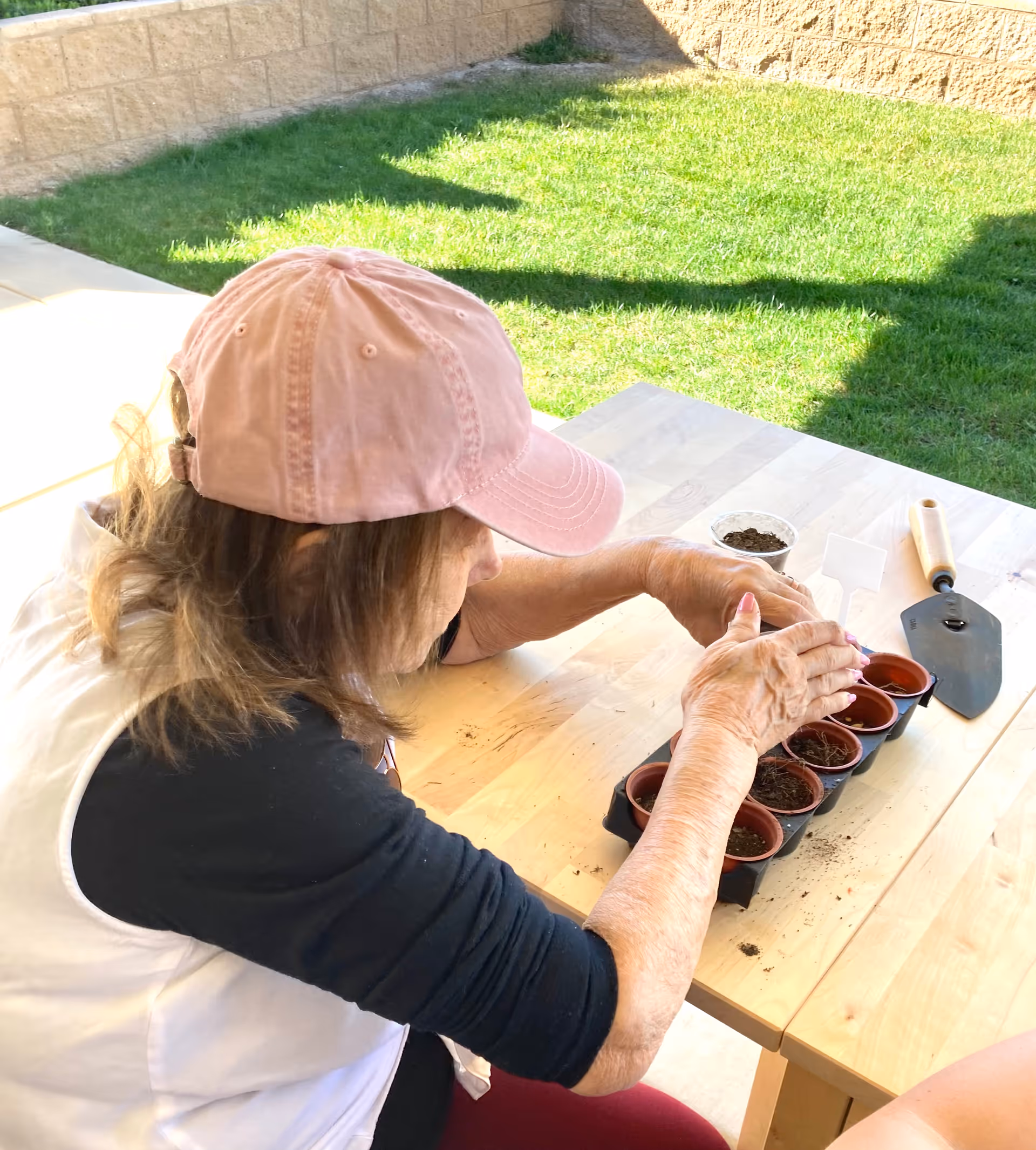 An elderly woman wearing a pink baseball cap and a white and black shirt is seated at a wooden table outdoors, planting seeds in small pots filled with soil. A gardening trowel and a small container of soil are also on the table. The background shows a grassy area and a stone wall.