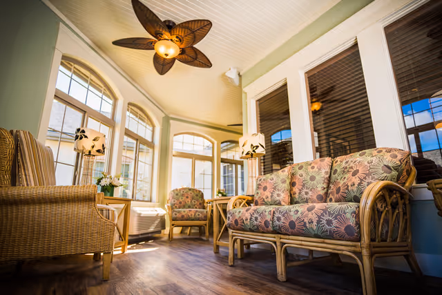 A bright and cozy sitting area with wicker furniture including a floral-patterned sofa and chairs, two side tables with floral lamps, large windows letting in natural light, and a ceiling fan with leaf-shaped blades.