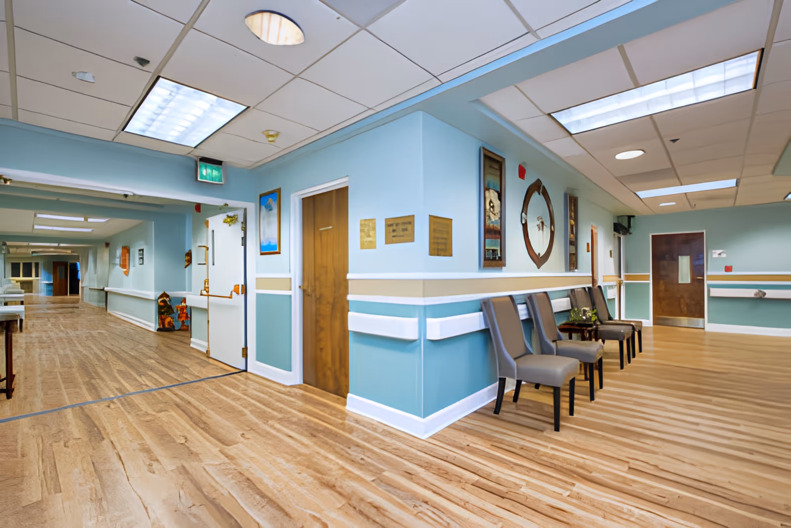 A wide, well-lit assisted living hallway with teal walls, a row of chairs, wooden doors, and hardwood floors.