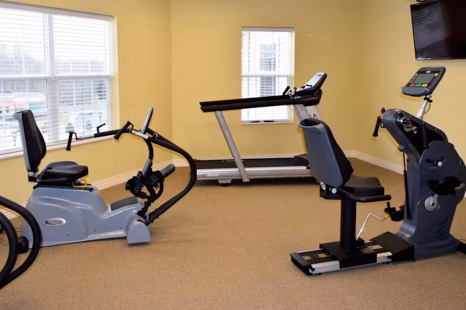 A small exercise room with beige walls and carpeted floor containing three pieces of exercise equipment: a recumbent bike, a treadmill, and a seated elliptical machine. The room has two windows with white blinds and a wall-mounted TV.