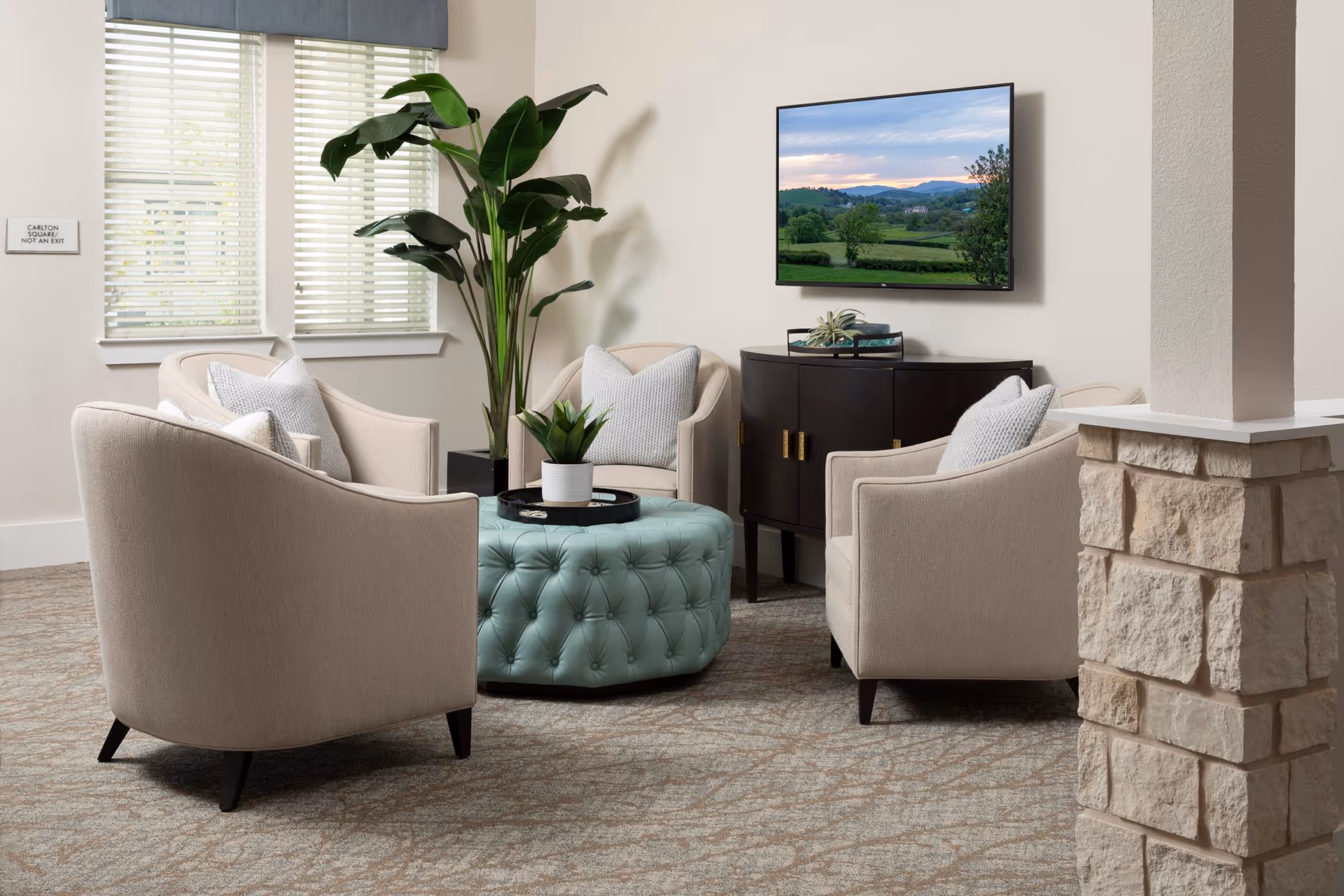 A cozy seating area in a senior living facility with four beige armchairs arranged around a round, tufted light blue ottoman. A small black tray with a potted plant sits on the ottoman. Behind the chairs, there is a dark wooden cabinet with decorative items on top and a flat-screen TV mounted on the wall displaying a scenic landscape. A large green plant is placed near two windows with white blinds, and a stone pillar is visible on the right side of the image.