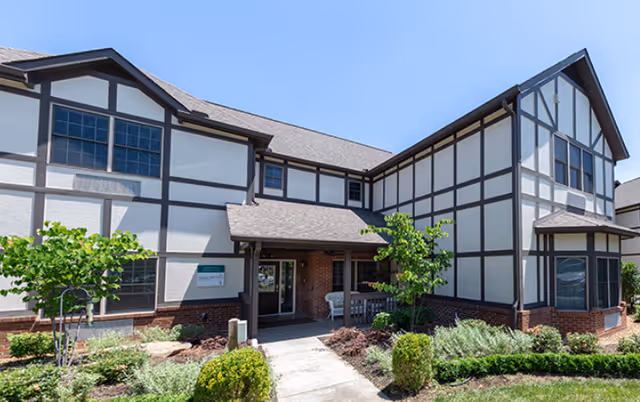 Exterior view of Deane Hill Place Senior Living facility showing a two-story building with Tudor-style architecture, white walls with dark wooden beams, a covered entrance, and landscaped greenery including bushes and small trees.
