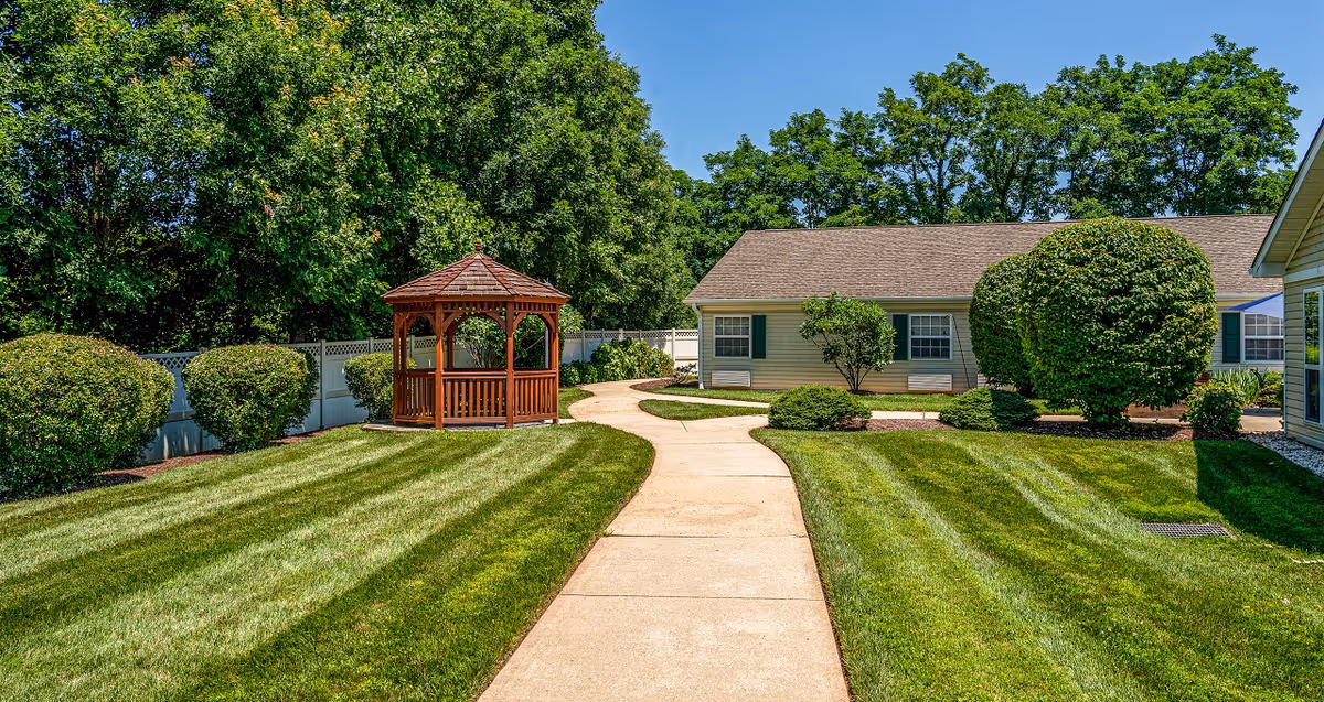 A well-maintained outdoor garden area with a concrete pathway leading to a wooden gazebo. The garden is surrounded by neatly trimmed bushes and trees, with a building visible in the background under a clear blue sky.