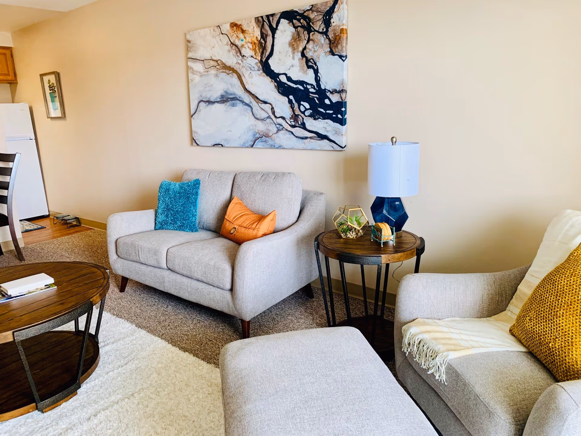 A cozy living room with a light gray loveseat and armchair, each adorned with colorful throw pillows. A wooden coffee table and side table with a blue lamp and decorative items are also visible. A large abstract painting hangs on the beige wall behind the loveseat. In the background, part of a dining area and a white refrigerator can be seen.