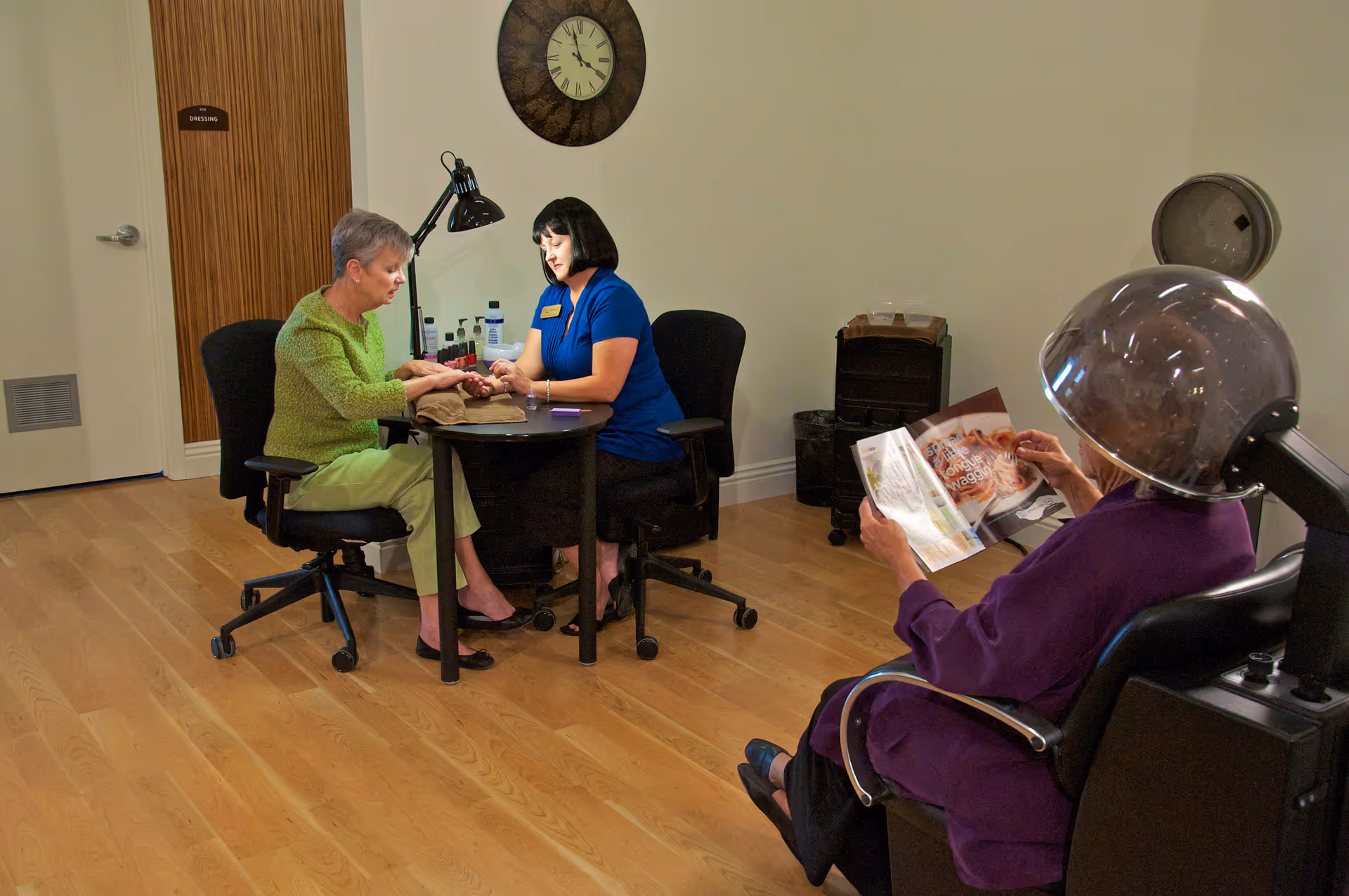 A room in a retirement community where a woman in a blue uniform is giving a manicure to an elderly woman wearing a green sweater. Another elderly woman in a purple robe is sitting under a hair dryer, reading a magazine. The room has wooden flooring, a wall clock, and a door labeled 'Dressing'.