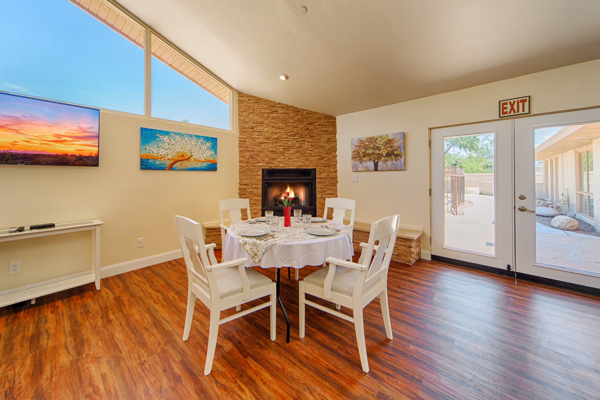 A cozy dining area with a round table set for four, featuring white chairs and a tablecloth. The room has a stone fireplace with a fire burning, colorful paintings on the walls, a large window letting in natural light, and glass doors leading outside. The floor is wood with a warm finish.