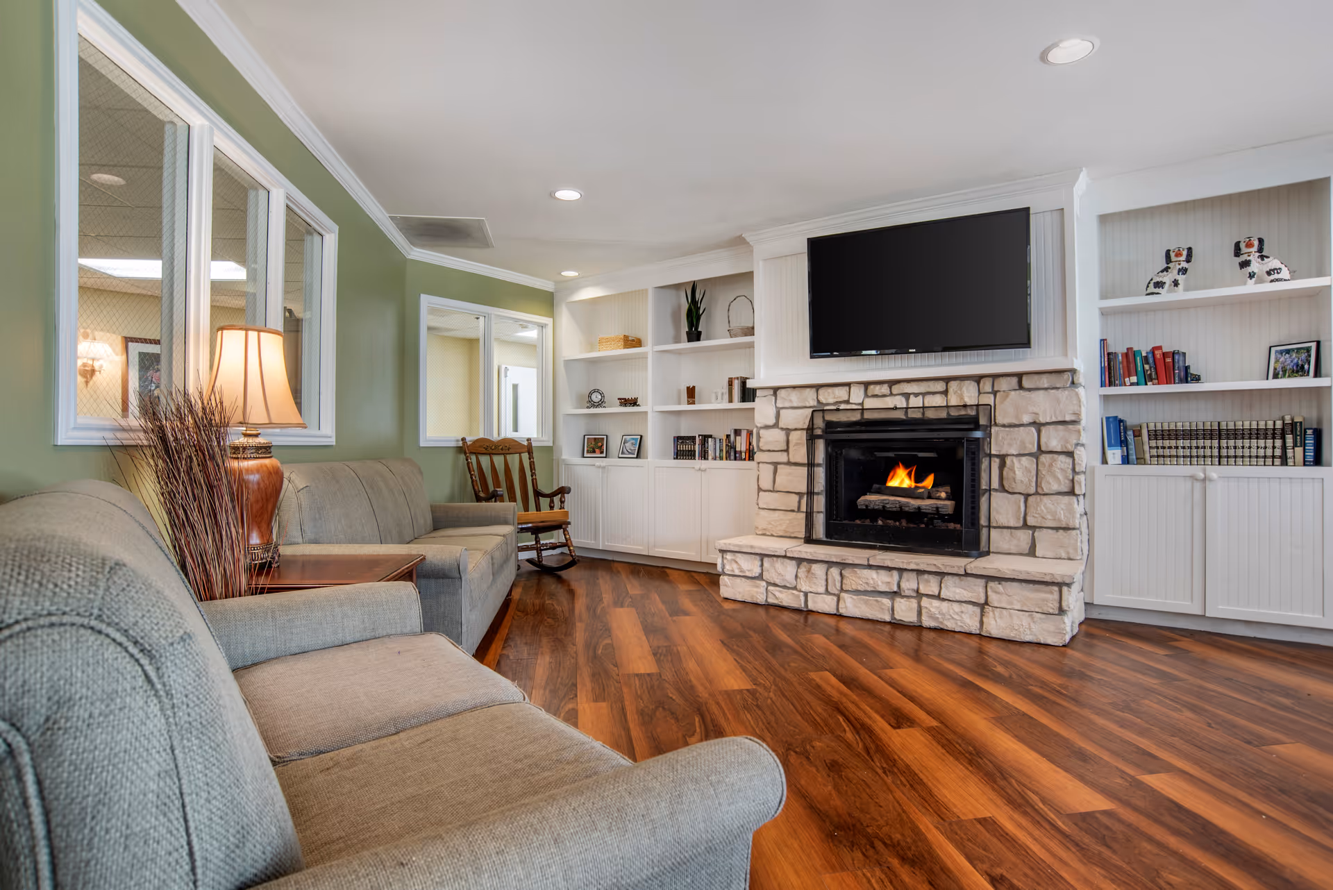 A cozy living room with a stone fireplace and a flat-screen TV mounted above it. The room features wooden flooring, two gray upholstered sofas, a wooden rocking chair, built-in white shelves with books and decorative items, a table lamp, and green painted walls with windows looking into another room.