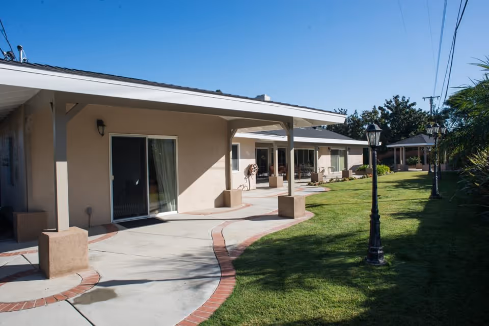 Outdoor view of a single-story assisted living facility with a covered walkway, sliding glass doors, green lawn, and vintage-style lamp posts under a clear blue sky.