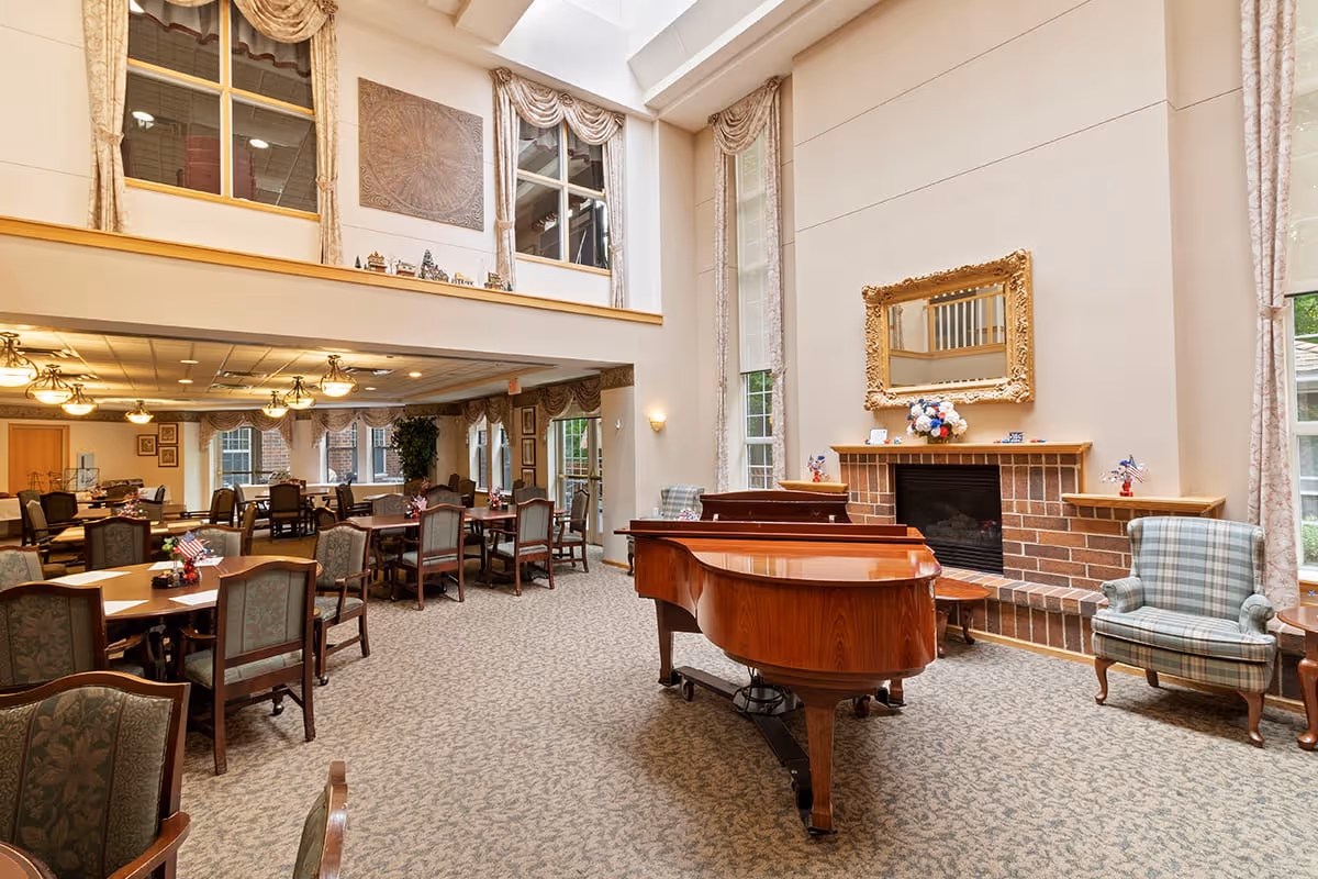 A spacious and elegant common area in Eagle Crest Assisted Living featuring a grand piano in the foreground, a brick fireplace with a gold-framed mirror above it, and several tables and chairs arranged for dining or socializing. The room has large windows with draped curtains allowing natural light to enter, and a high ceiling with a mezzanine level visible.