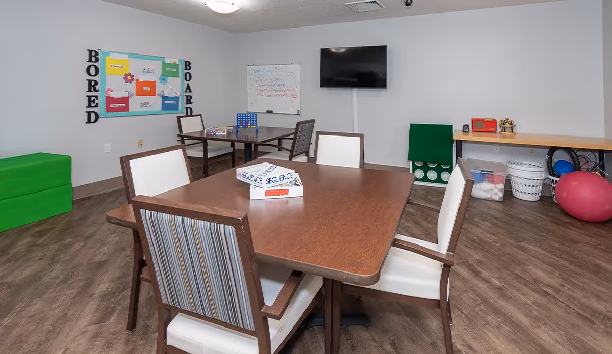 A senior living facility activity room with a wooden table and six chairs. On the table are board games including Sequence and Sorry. The room has a bulletin board labeled 'Bored Board', a whiteboard with writing, a wall-mounted TV, and various recreational items like a large green Connect Four game, a red radio, baskets, and exercise balls. The floor is wood, and the walls are light gray.