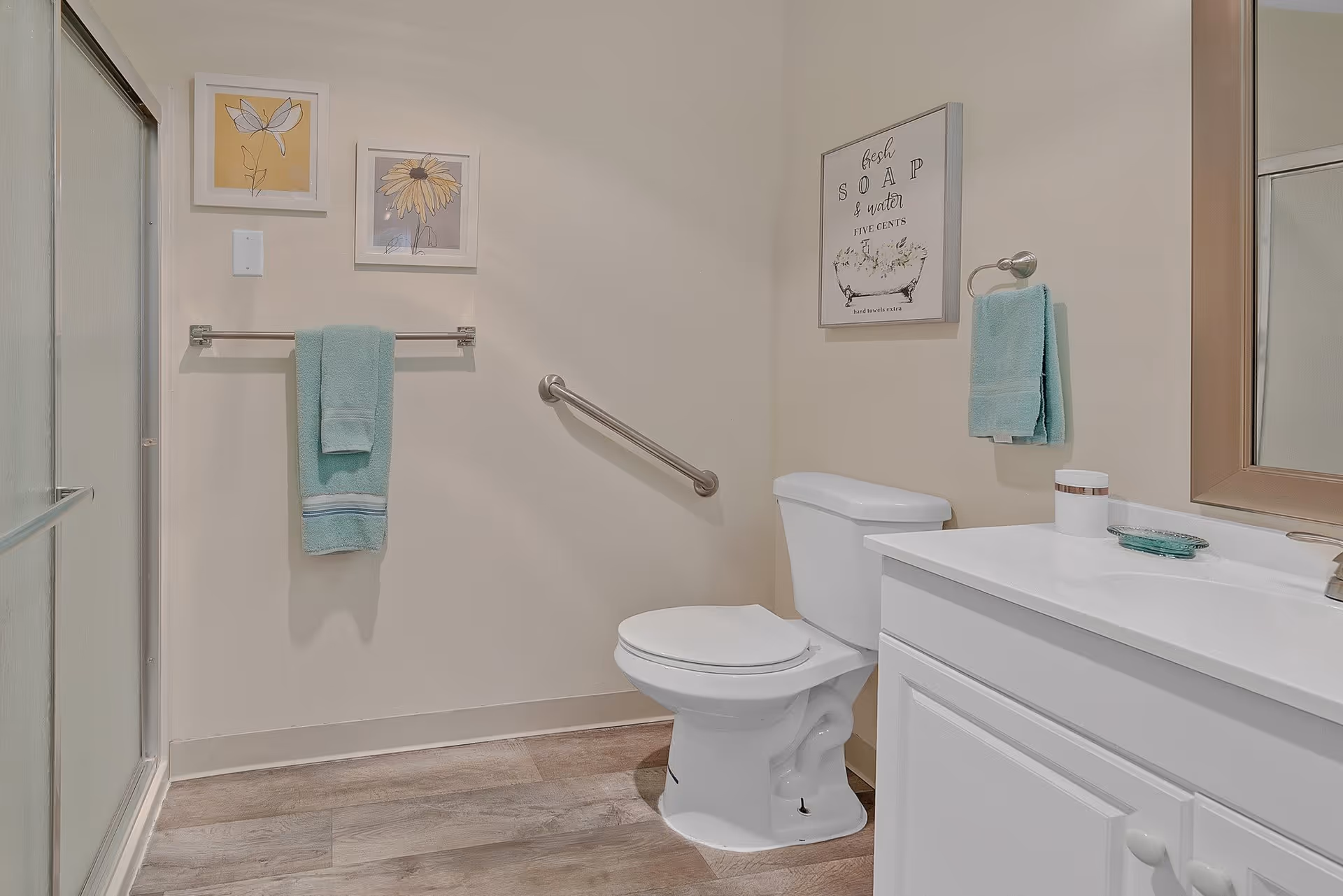A clean bathroom with a white toilet, white vanity with a sink, and a large mirror. There are two light blue towels hanging on towel racks, one near the toilet and one on the wall. The bathroom has light-colored walls and wood-style flooring. Two framed floral pictures hang on the wall above one towel rack, and a framed sign with the text 'fresh SOAP & water FIVE CENTS' hangs above the toilet. A metal grab bar is installed on the wall next to the toilet. A shower with frosted glass doors is visible on the left side.