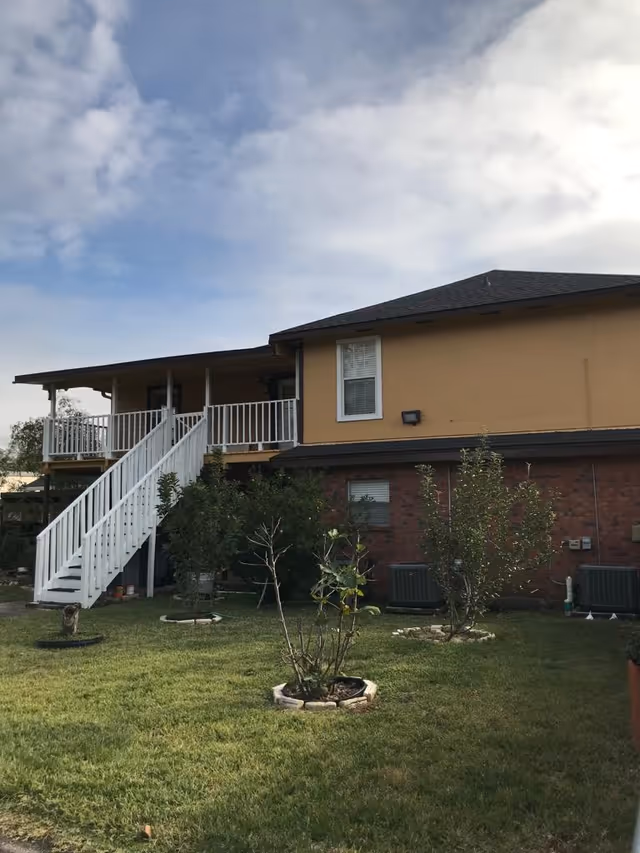 View of the exterior of a two-story building with a yellow upper level and brick lower level. There is a white staircase leading to a covered porch on the upper floor. The foreground shows a grassy yard with small plants and shrubs. The sky is partly cloudy.