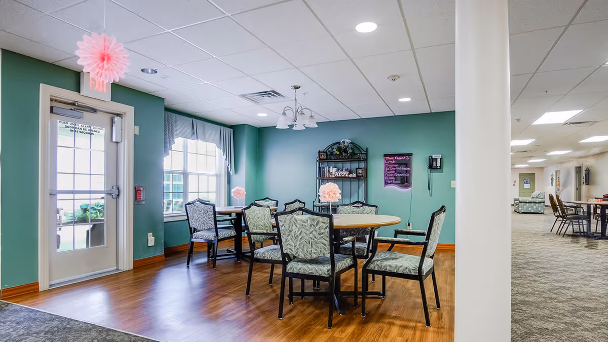 A dining area in an assisted living facility with round tables and cushioned chairs arranged around them. The walls are painted green, and there is a window with curtains letting in natural light. A door with an exit sign above it is visible on the left. A black shelving unit with decorative items and a menu board are on the far wall. The floor is a mix of wood and carpet, and the ceiling has recessed lighting.