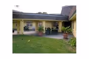 Outdoor patio area at Pine Trail Nursing and Rehab Center with a covered seating area featuring several tables and chairs, potted plants, and a grassy lawn in the foreground.