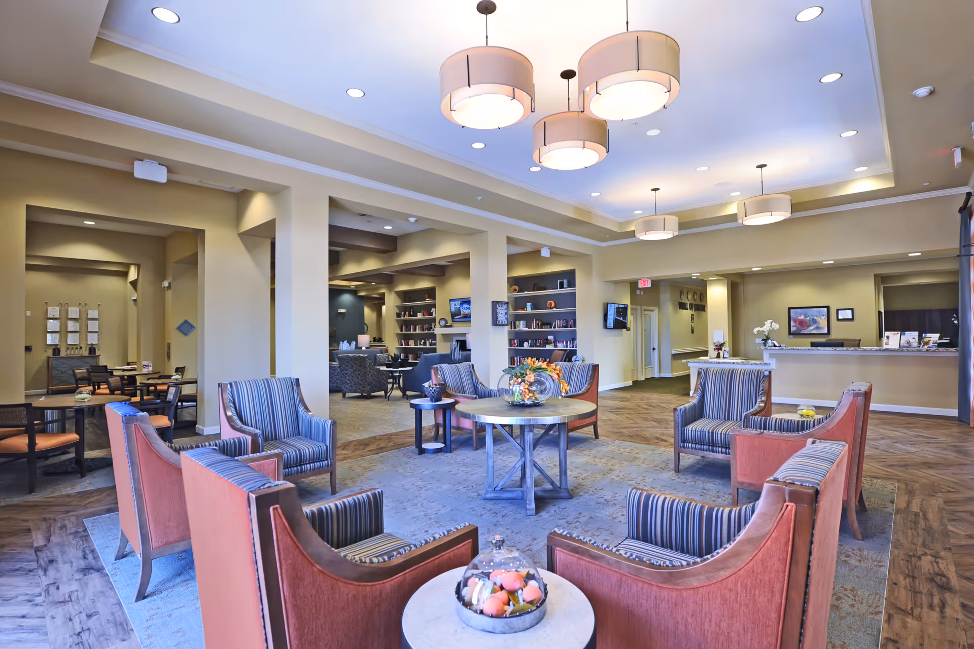 A spacious senior living facility common area with a circular seating arrangement of striped and red upholstered chairs around a central round table with a floral centerpiece. The room features warm beige walls, wooden flooring, built-in bookshelves, multiple pendant lights hanging from a tray ceiling, and a reception desk in the background.