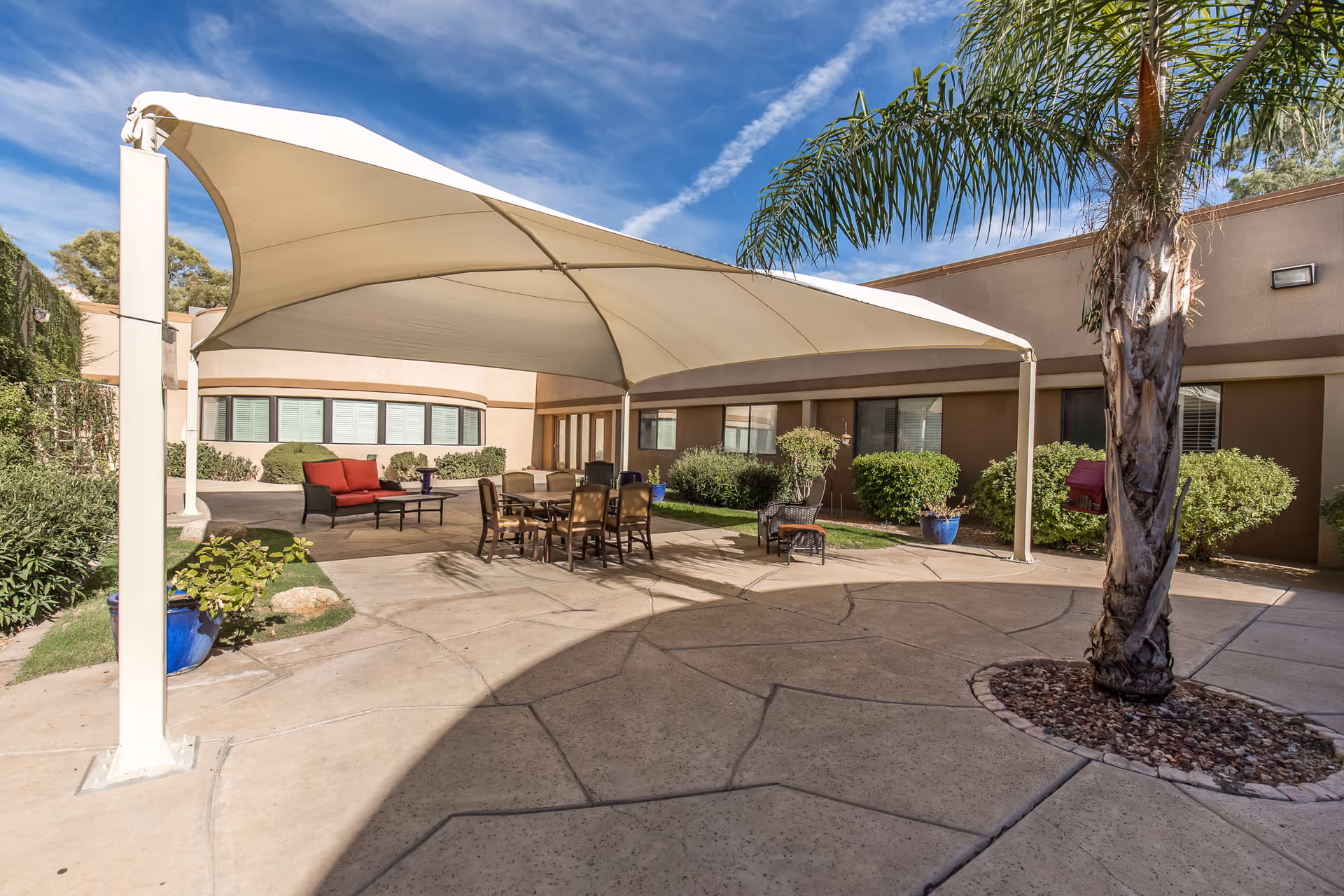 Outdoor patio area at Ridgecrest Post Acute with a large white canopy providing shade over a seating area that includes a table with chairs and a red cushioned bench. The patio is surrounded by bushes, potted plants, and a palm tree, with a building in the background under a blue sky with some clouds.