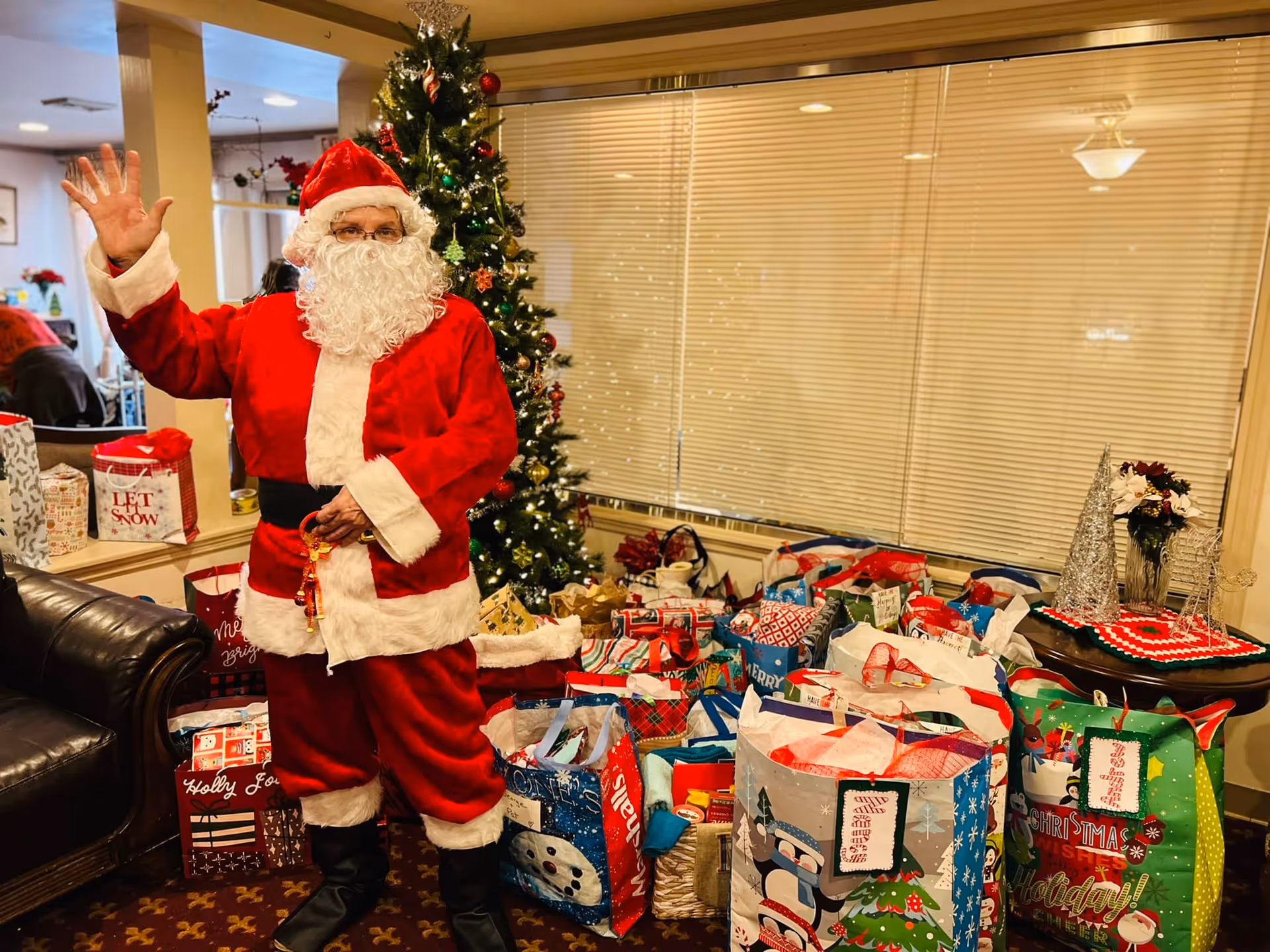 A person dressed as Santa Claus waving while standing next to a Christmas tree and a large collection of colorful holiday gift bags in a cozy indoor setting with a leather couch and window blinds in the background.