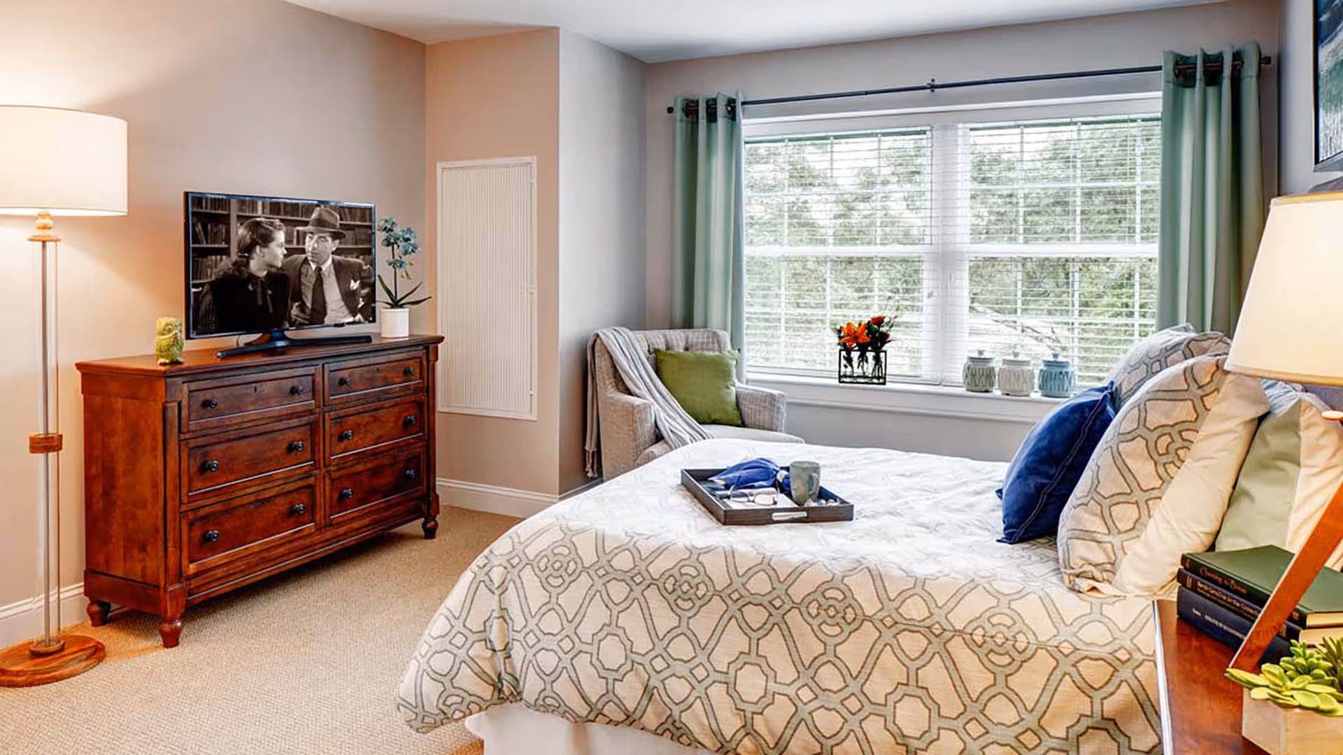 A cozy bedroom with a large window covered by light green curtains, a bed with patterned bedding and multiple pillows, a wooden dresser with a TV showing a black and white movie, a floor lamp, an armchair with a green pillow and a throw blanket, and decorative items on the windowsill and nightstand.