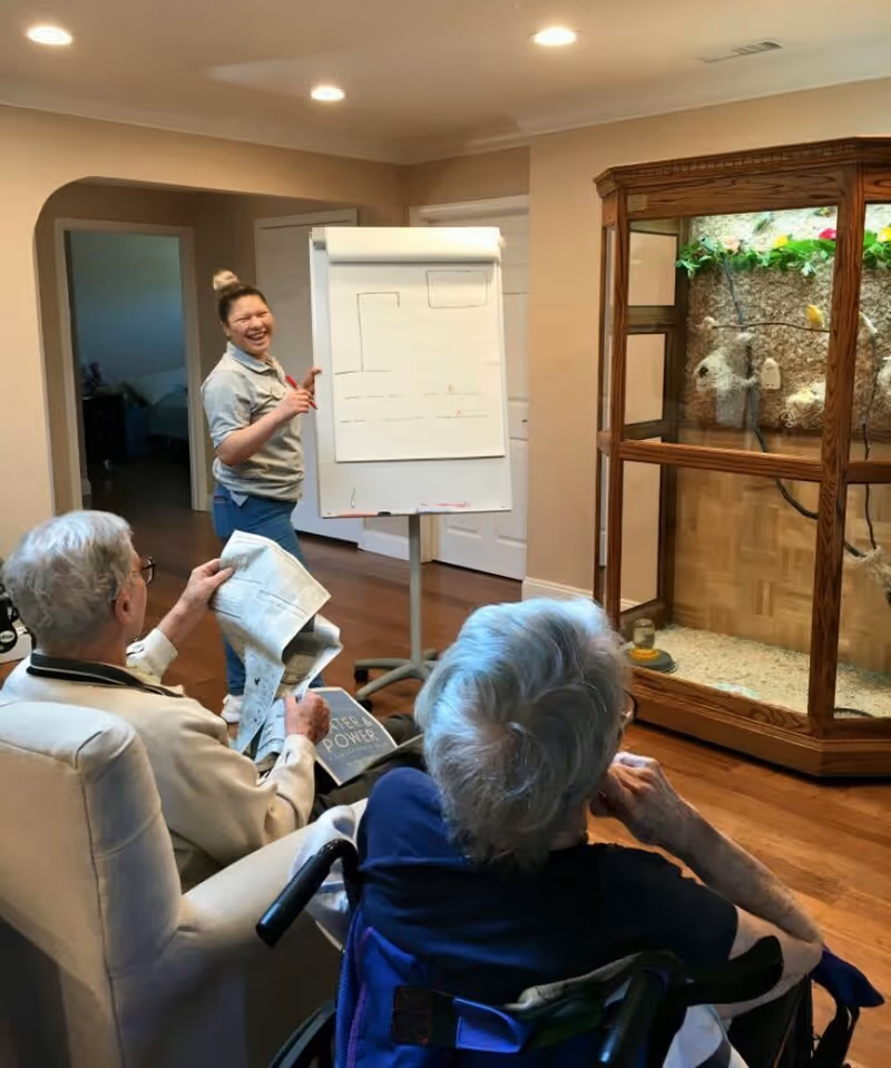 A caregiver stands next to a flip chart with simple drawings, smiling and engaging with two elderly individuals seated in a living room. One elderly person is in a wheelchair, and the other is holding a newspaper. The room has wooden flooring and a large wooden birdcage with birds inside.