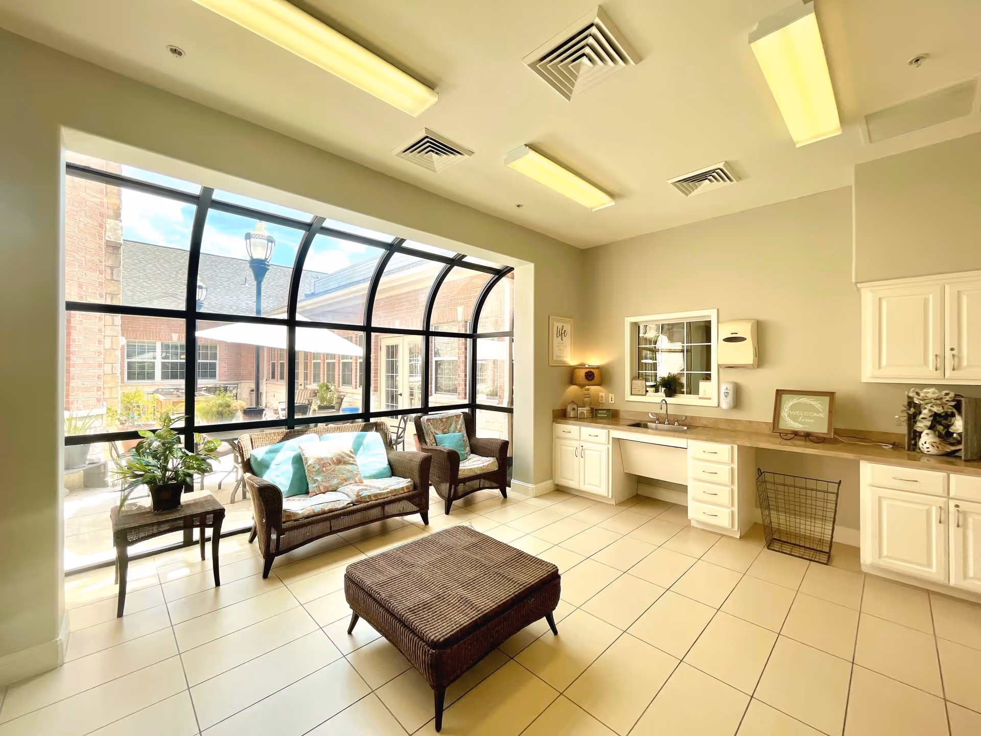 A bright, clean room with large curved windows overlooking an outdoor patio. Inside, there is a wicker loveseat and chair with cushions, a wicker ottoman, a small side table with a plant, and a long countertop with cabinets, a sink, and a paper towel dispenser. The room has tiled floors and fluorescent ceiling lights.