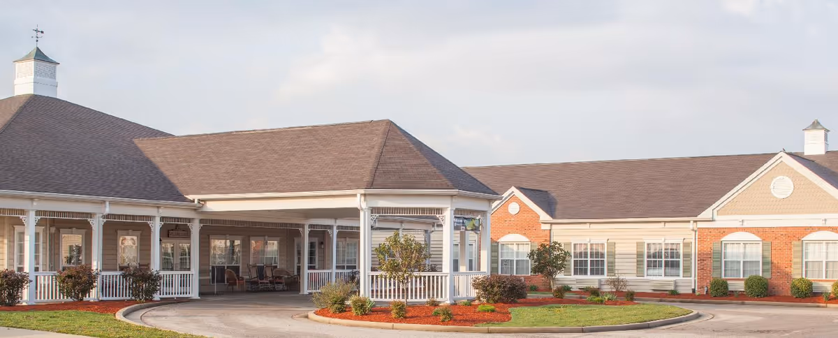 Exterior view of a single-story senior living facility building with a covered entrance, white railings, and landscaped bushes and trees around the driveway.