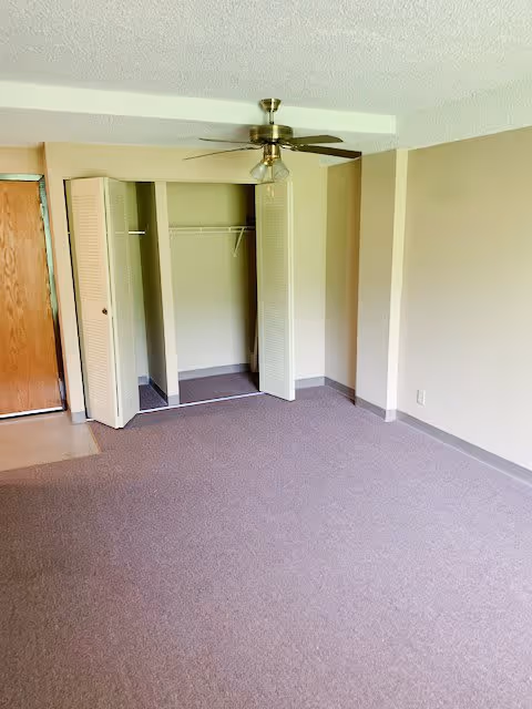 Empty carpeted room with an open bi-fold closet, ceiling fan, and a wooden entry door.