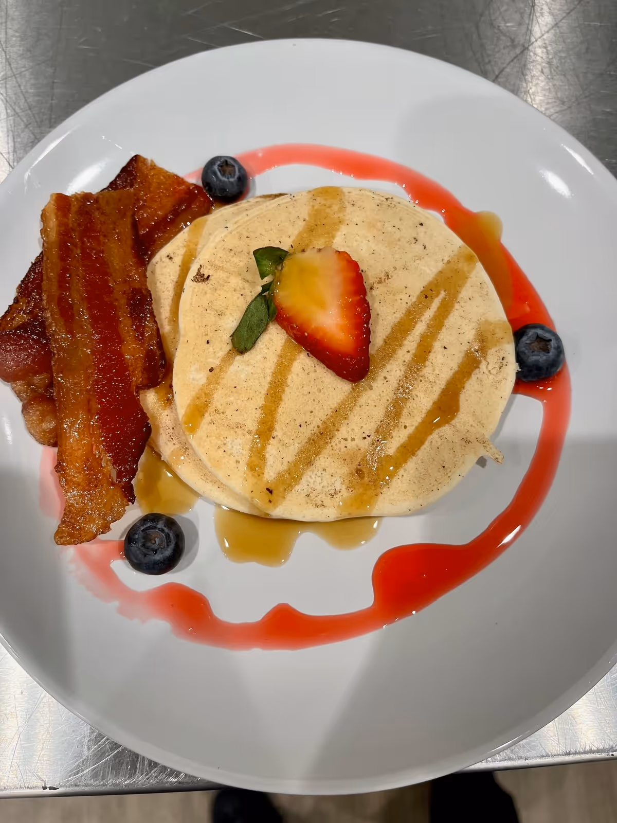 A white plate with two pancakes drizzled with syrup and topped with a halved strawberry. There are two strips of crispy bacon on the side and a few blueberries scattered around the plate with a red sauce decoration.