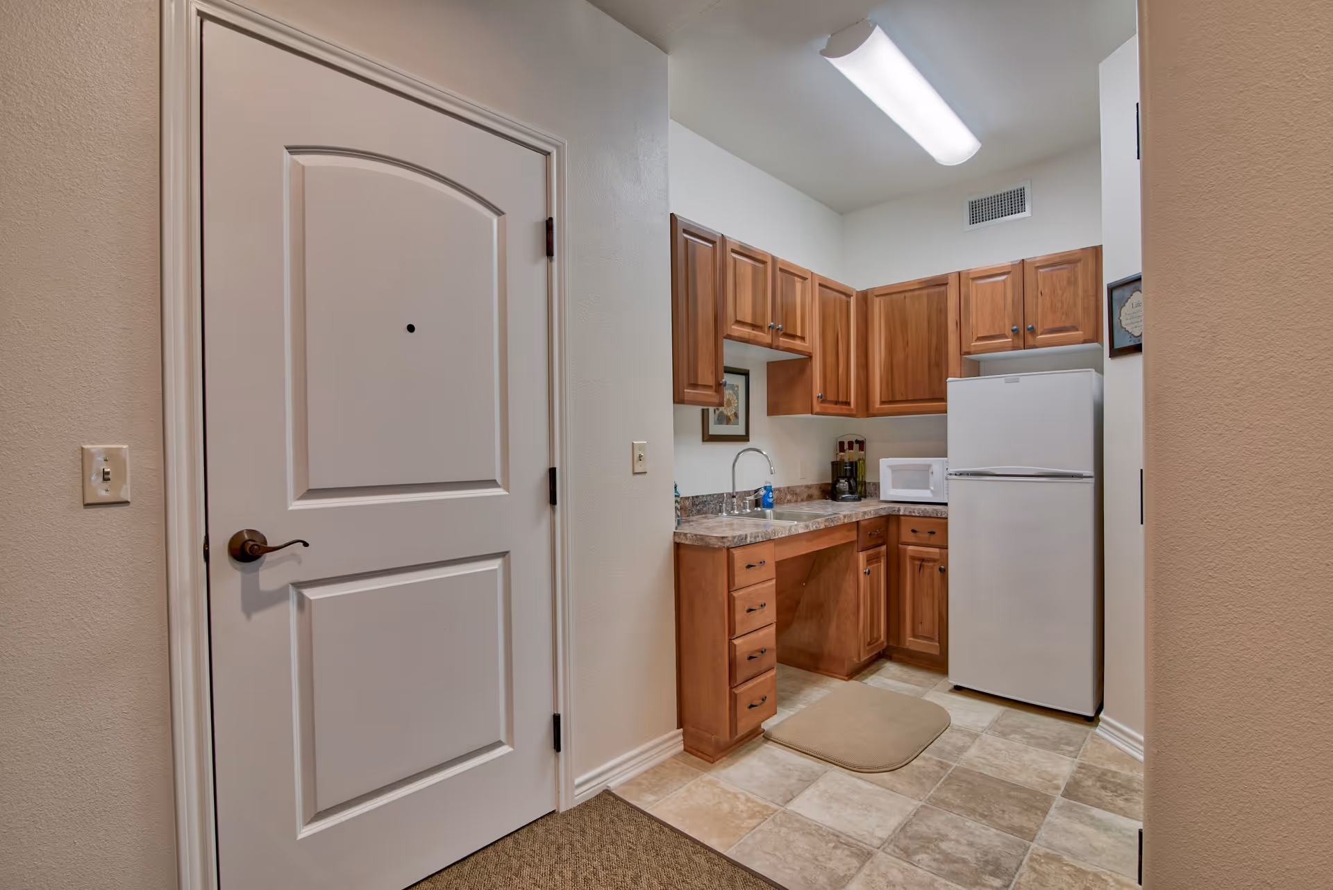 Small kitchen area with wooden cabinets, a white refrigerator, a microwave, a sink, and a countertop. The floor has beige and light brown tiles, and there is a white door with a peephole on the left side of the image.