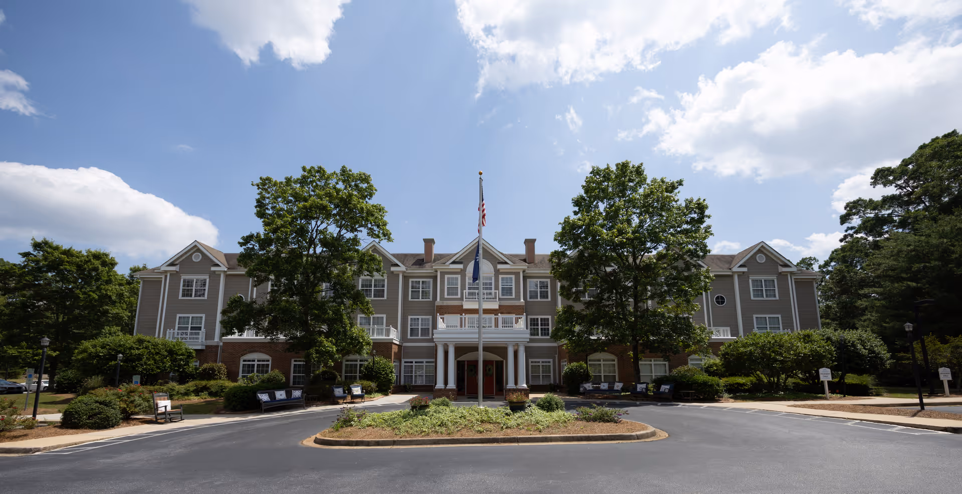 Front exterior view of a large, three-story senior living facility building named The Gables, with a circular driveway, landscaped greenery, benches, and two large trees flanking the entrance under a partly cloudy sky.