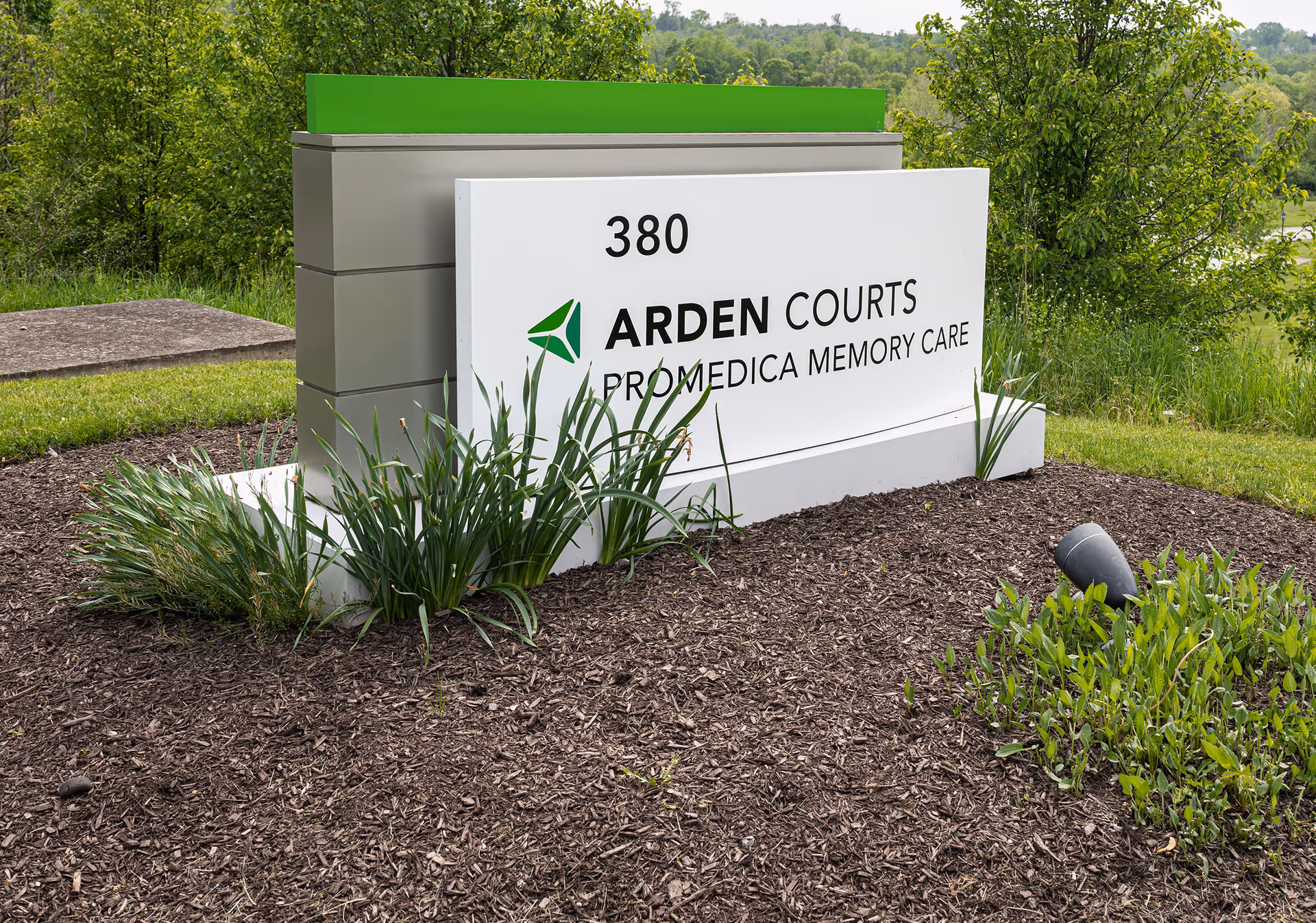 Outdoor sign for Arden Courts ProMedica Memory Care Community at 380, surrounded by mulch and green plants with trees and grass in the background.