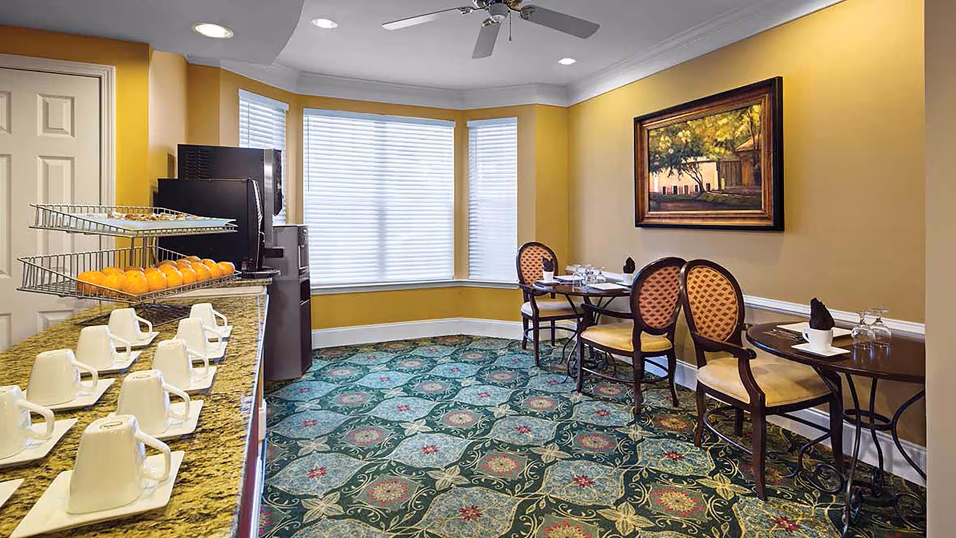A cozy dining area with two small round tables, each set with chairs and tableware. The room has yellow walls, a patterned green carpet, and a large window with white blinds. A countertop with white cups and saucers, a tiered fruit basket with oranges, and a refrigerator are visible on the left side. A framed painting hangs on the wall.