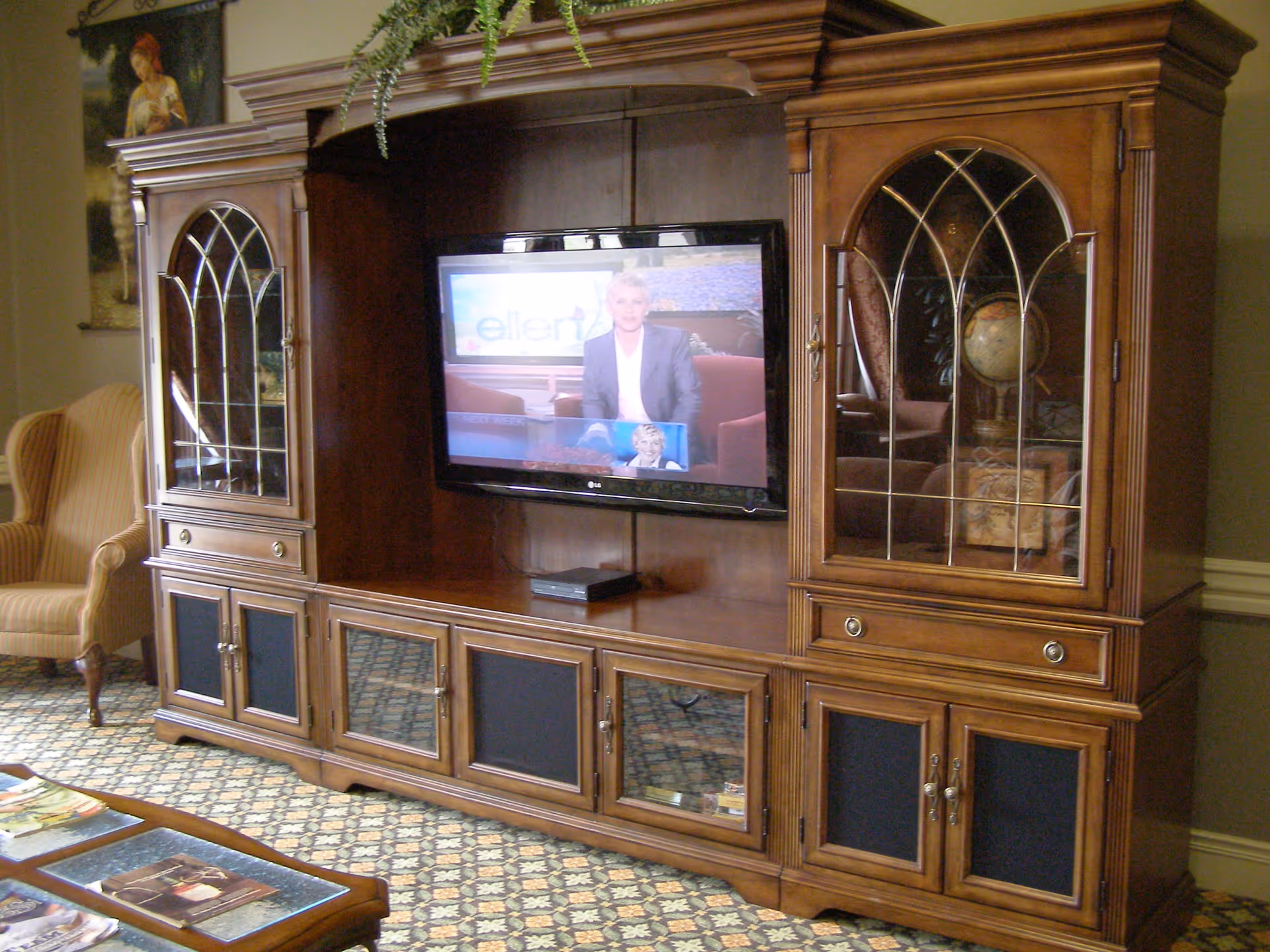 A wooden entertainment center with glass-paneled cabinets on each side and a flat-screen TV in the middle displaying a talk show. To the left, there is a striped upholstered armchair and a framed painting on the wall. The floor is covered with a patterned carpet.