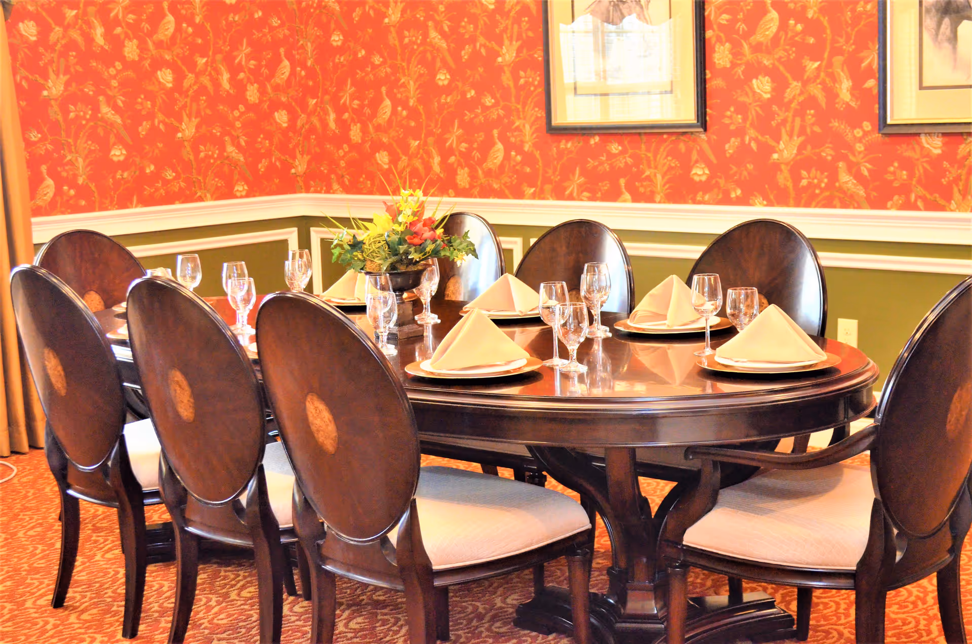 A formal dining room with a large oval wooden table set for eight people. Each place setting includes a white plate, a folded beige napkin, and multiple wine glasses. The room features red wallpaper with a gold floral pattern, green wainscoting, and framed artwork on the walls. A floral centerpiece decorates the middle of the table.