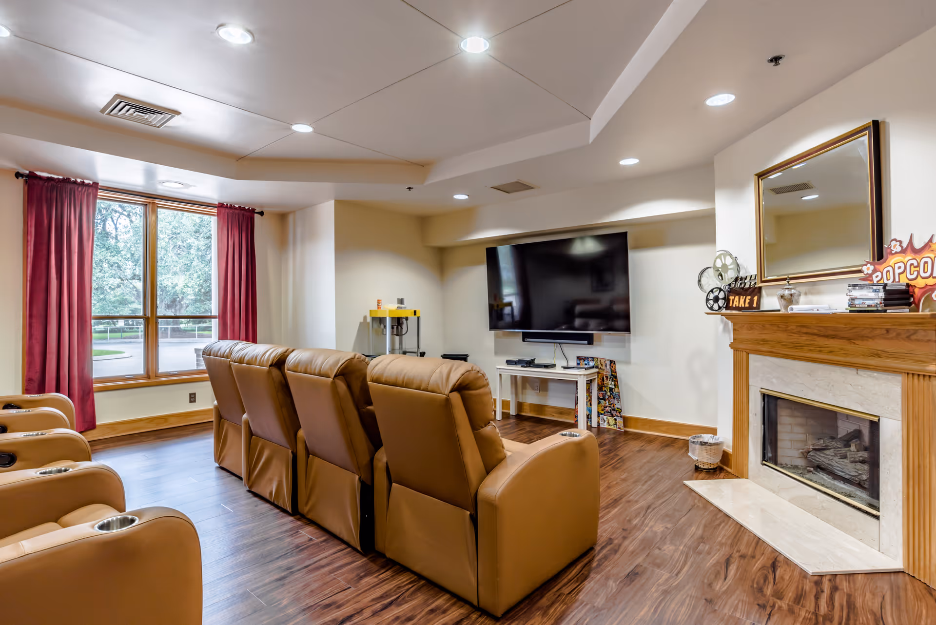 A cozy living room with a row of six tan leather recliners facing a large flat-screen TV mounted on the wall. The room features wood flooring, a fireplace with a wooden mantle decorated with a mirror, film reels, and a popcorn sign. A large window with red curtains allows natural light to enter the space.