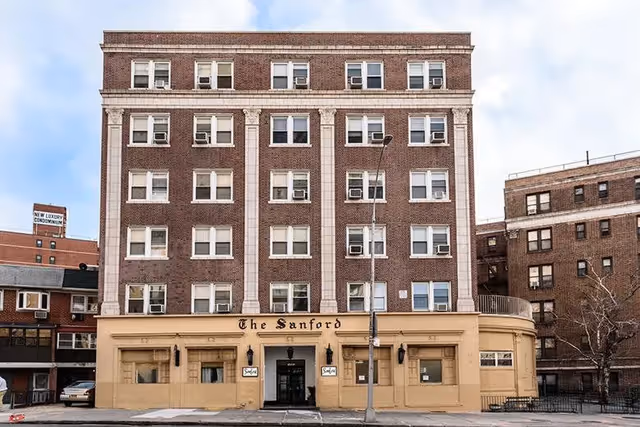 Front exterior view of a multi-story brick building named The Sanford with several windows and air conditioning units. The lower part of the building is painted beige with the name displayed above the entrance. A streetlamp and sidewalk are visible in front of the building.