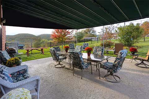 Outdoor covered patio area with a glass-top table surrounded by eight cushioned chairs with floral patterns. There are potted flowers on the table and additional seating including a white wicker chair with cushions. The patio overlooks a fenced grassy area with trees showing autumn colors and hills in the background under a cloudy sky.
