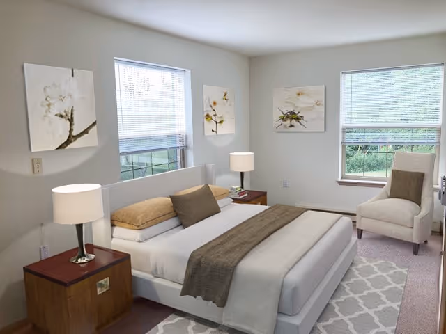 A bright bedroom with a large bed featuring white and beige bedding, two wooden nightstands with lamps, a beige armchair near a window, and three floral paintings on the walls. The room has two windows with blinds partially open, allowing natural light to fill the space.