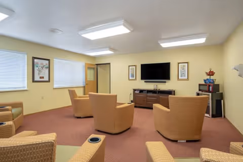 A cozy living room area in a senior living facility with several cushioned armchairs arranged facing a wall-mounted flat-screen TV. The room has beige walls, carpeted floor, two windows with blinds, framed artwork on the walls, and a wooden cabinet under the TV. There is also a small table with a coffee maker and supplies in the corner.
