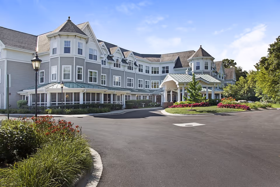 A large three-story senior living building with a covered entrance, circular driveway and landscaped flower beds under a blue sky.