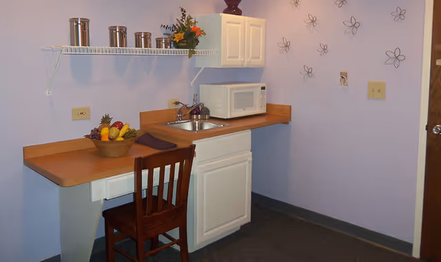 Small kitchenette with a countertop, sink, microwave, wooden chair, shelving with canisters and a bowl of fruit against a lavender wall with decorative wall flowers.