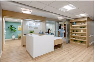 Bright wellness center reception with a white front desk, a seated receptionist, a staff member walking through a doorway, seating area, and wooden shelving with plants.