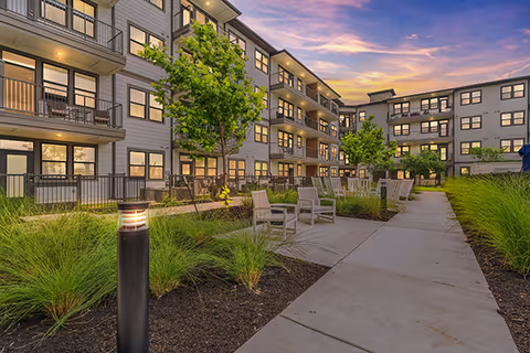Courtyard with a paved walkway, benches, landscaping, and a multi-story apartment building with balconies at dusk.