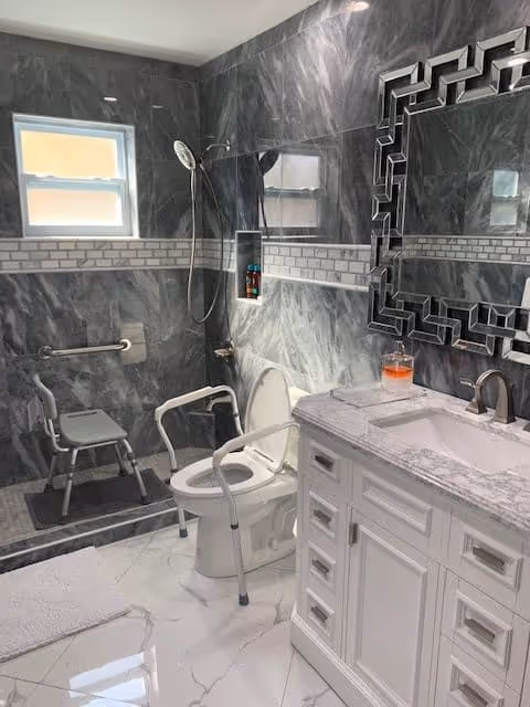 A modern bathroom with gray marble walls and white tiled floor. The bathroom features a walk-in shower with a handheld showerhead and a shower chair. There is a toilet with safety rails around it. A white vanity with a marble countertop and a decorative mirror above it is visible. A small window provides natural light.