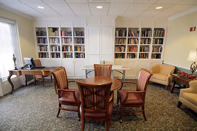 A cozy common area with a round wooden table surrounded by four upholstered chairs in the center. Behind the table is a wall with built-in white bookshelves filled with books. To the left, there is a wooden desk with a computer and chair near a window with blinds. On the right side, there are two beige upholstered armchairs and a small wooden side table with a lamp and decorative items. The room has patterned carpet and soft yellow walls.