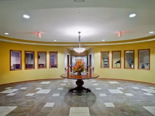A spacious circular interior room with a tiled floor featuring a mix of brown and beige tiles. In the center is a round wooden table with a floral arrangement. The walls are painted yellow and have several rectangular windows looking into adjacent rooms. A chandelier hangs from the ceiling above the table.