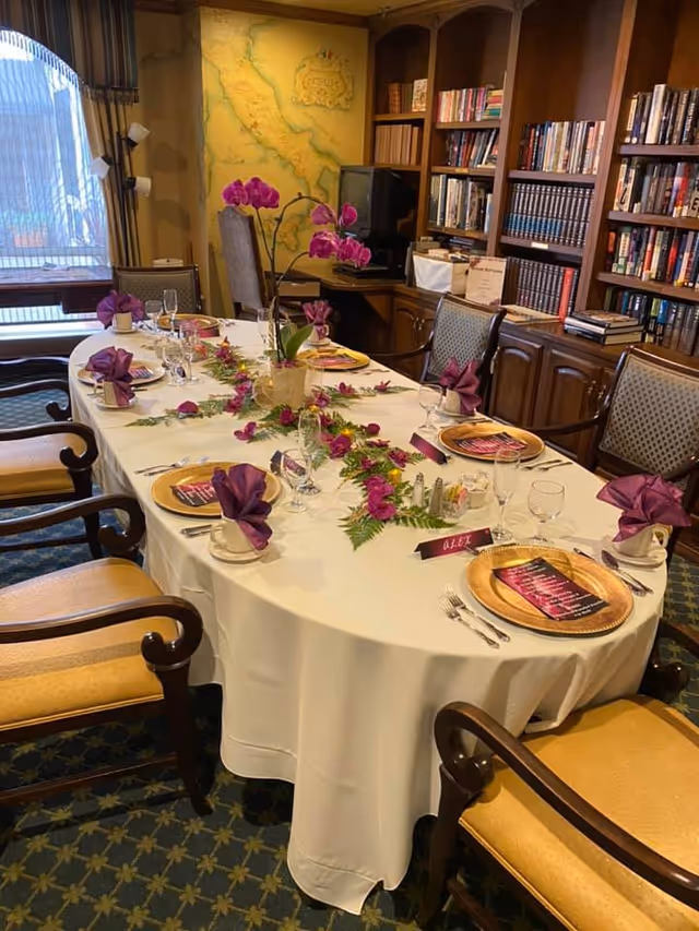 A formal dining room with a long oval table covered with a white tablecloth, set with gold charger plates, purple folded napkins, glassware, and silverware. The table is decorated with a centerpiece of purple orchids and green foliage. The room has wooden bookshelves filled with books, a large map on the wall, and upholstered chairs around the table.