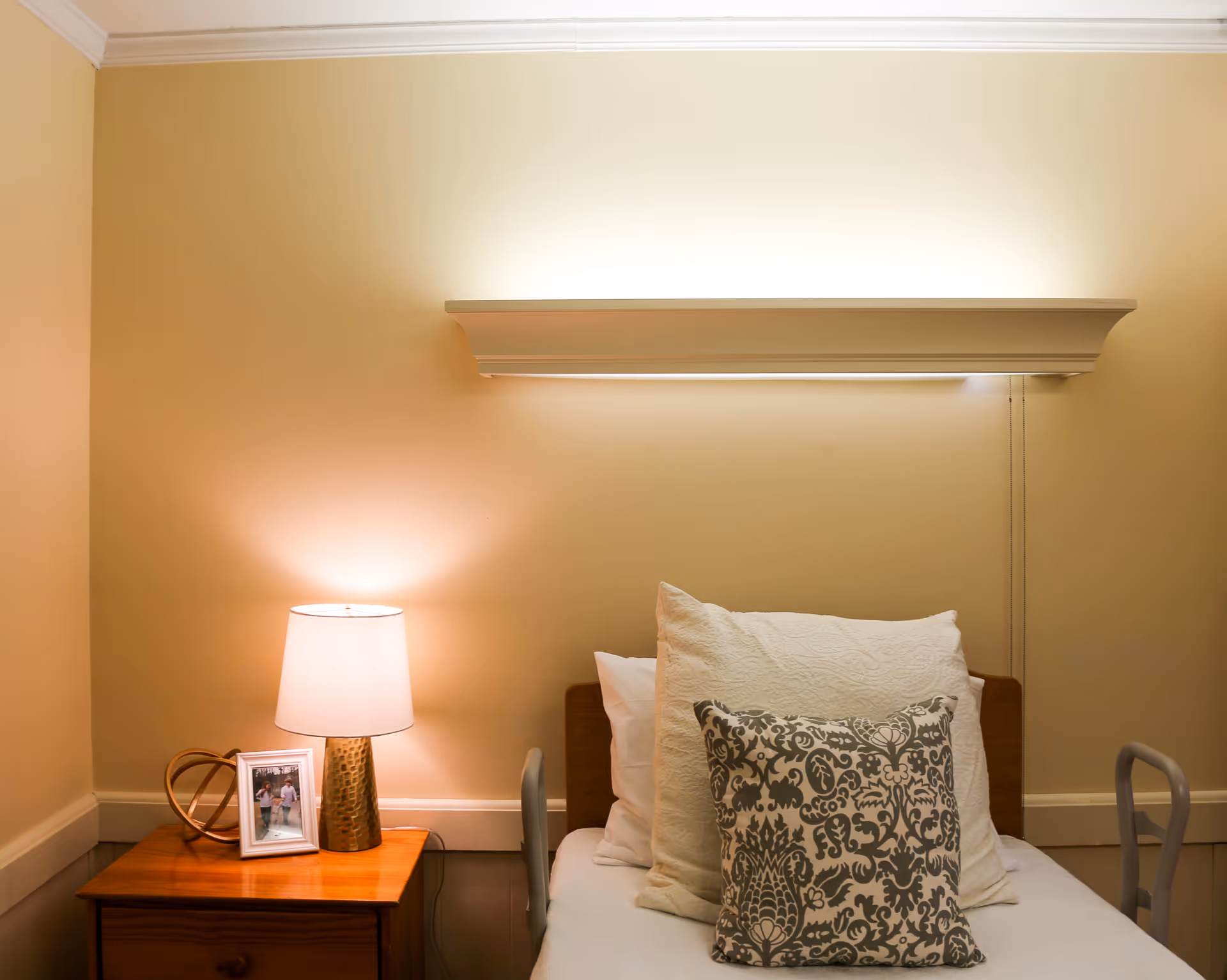 A cozy bedroom corner in a senior living facility featuring a single bed with multiple pillows, including a decorative patterned pillow. Next to the bed is a wooden nightstand with a lit table lamp, a framed photo, and a decorative item. The walls are painted a soft beige color, and there is a wall-mounted light fixture above the bed providing additional illumination.